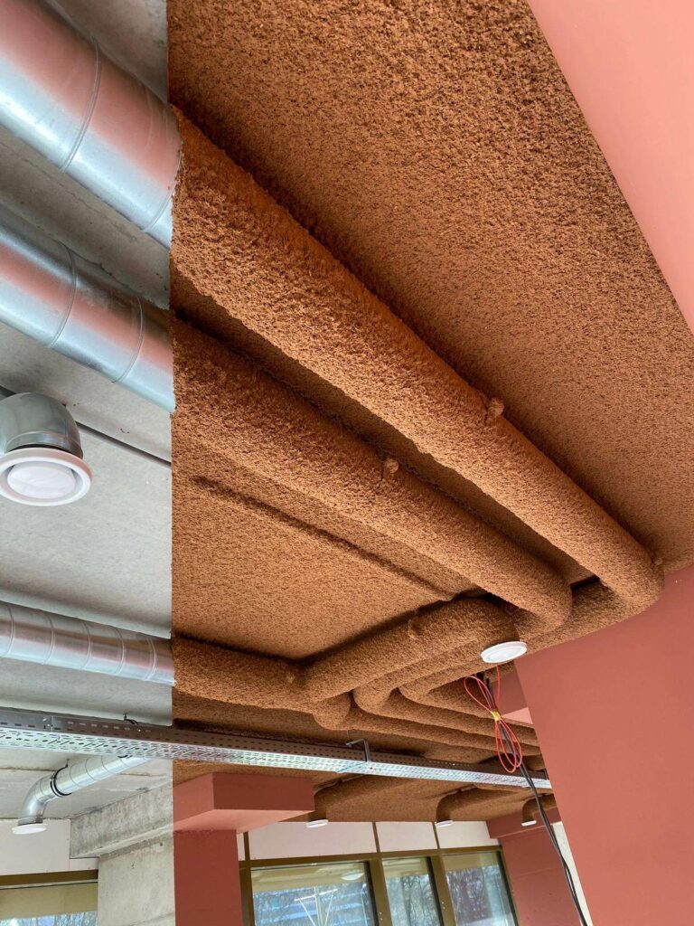 Sous-face de poutres béton traitée en projection acoustique terracotta, à côté d'une zone non traitée aux gaines apparentes