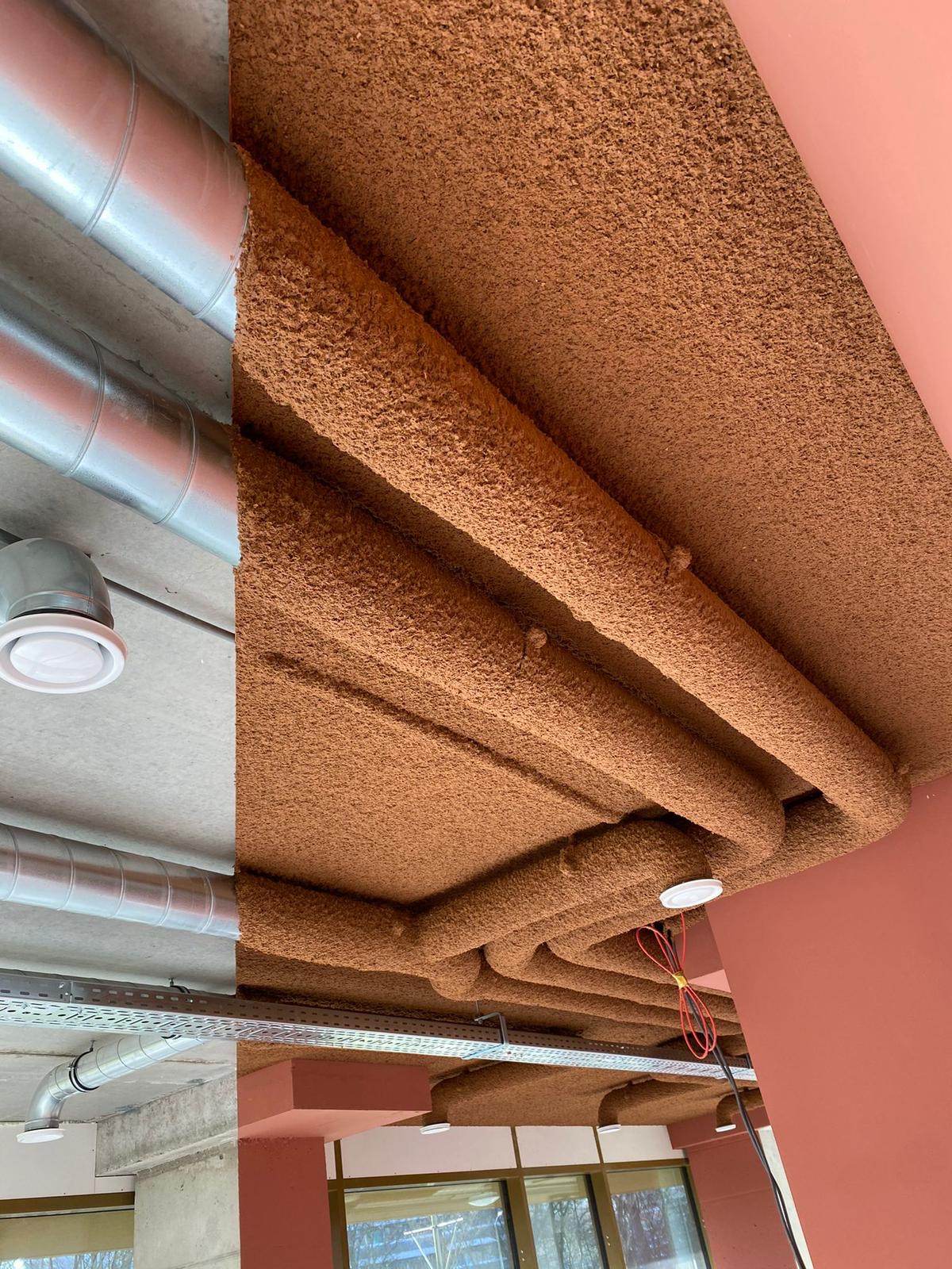 Sous-face de poutres béton traitée en projection acoustique terracotta, à côté d'une zone non traitée aux gaines apparentes